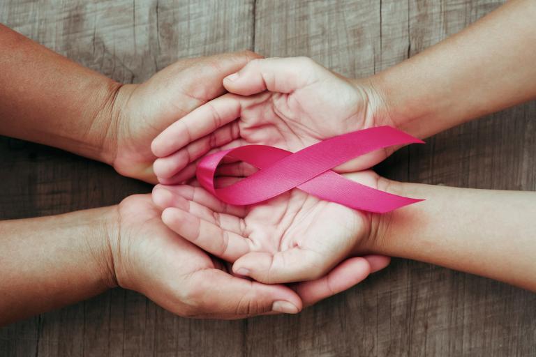 A larger pair of hands cupping a smaller pair of hands palms up with a pink breast cancer awareness ribbon.